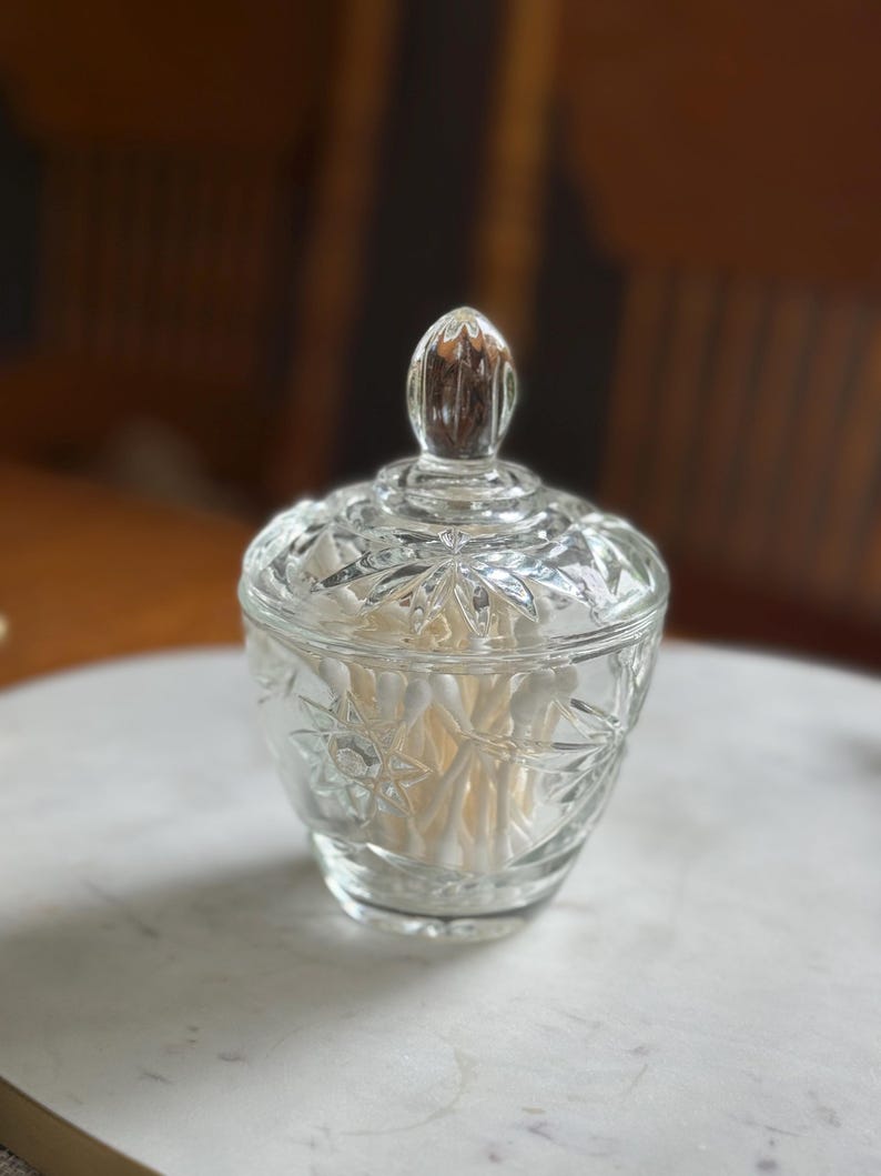 May include: A clear glass lidded jar featuring a faceted starburst pattern on the body and lid. The jar has a rounded base and a pointed finial handle on top. Inside the container, a collection of white cotton swabs is clearly visible.