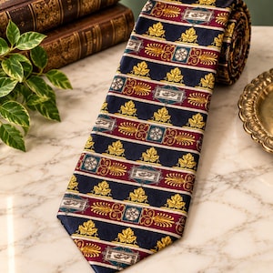 May include: A necktie featuring a repeating pattern of gold maple leaves on a dark navy blue background, interspersed with horizontal bands of deep burgundy, teal squares with white floral motifs, and rectangular silver-toned emblems.