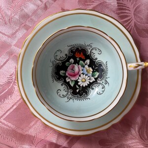 May include: A pale mint green teacup and saucer set featuring a central black medallion with a colorful floral bouquet of pink roses and white daisies. The rim of the cup and saucer is accented with a thin gold band and scalloped edges.