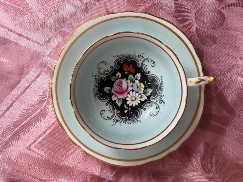 May include: A pale mint green teacup and saucer set featuring a central black medallion with a colorful floral bouquet of pink roses and white daisies. The rim of the cup and saucer is accented with a thin gold band and scalloped edges.