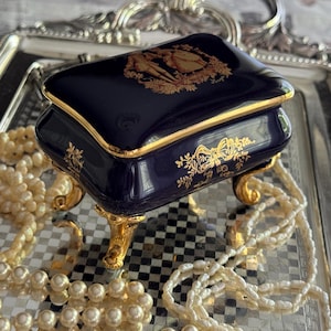 May include: A navy blue porcelain jewelry box with gold trim and ornate gold detailing. The box sits on gold feet and features a decorative design on the lid. A strand of pearls is draped around the box, resting on a silver tray.