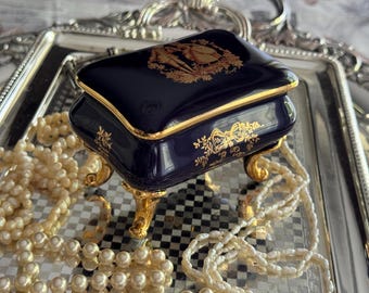 May include: A navy blue porcelain jewelry box with gold trim and ornate gold detailing. The box sits on gold feet and features a decorative design on the lid. A strand of pearls is draped around the box, resting on a silver tray.