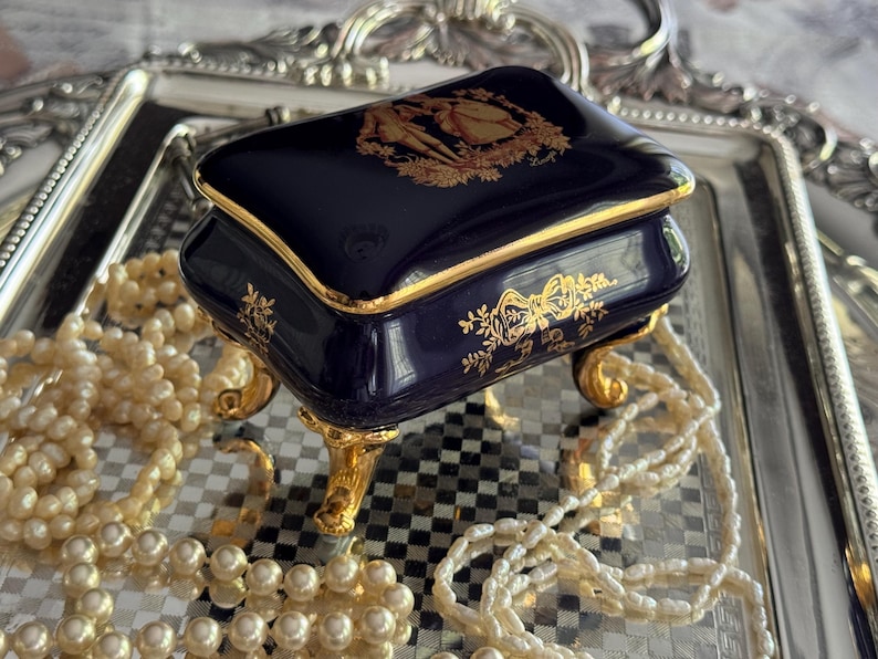 May include: A navy blue porcelain jewelry box with gold trim and ornate gold detailing. The box sits on gold feet and features a decorative design on the lid. A strand of pearls is draped around the box, resting on a silver tray.