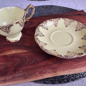 May include: A yellow teacup and saucer set with gold floral designs. The cup has a handle and the saucer has a scalloped edge.