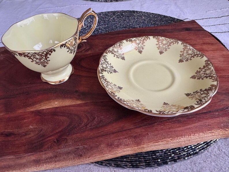 May include: A yellow teacup and saucer set with gold floral designs. The cup has a handle and the saucer has a scalloped edge.