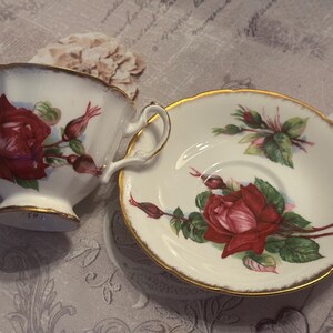 May include: A white teacup and saucer set with a red rose and bud design. The cup and saucer have gold trim. The teacup has a handle and sits next to the saucer on a patterned surface.