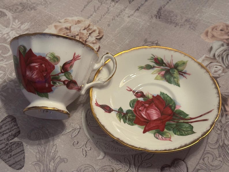 May include: A white teacup and saucer set with a red rose and bud design. The cup and saucer have gold trim. The teacup has a handle and sits next to the saucer on a patterned surface.