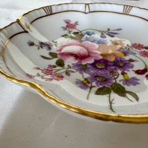 May include: A white porcelain dish with a scalloped edge and gold trim. The dish is decorated with a floral arrangement of pink roses, purple daisies, and other colorful flowers. A decorative piece for serving or display.