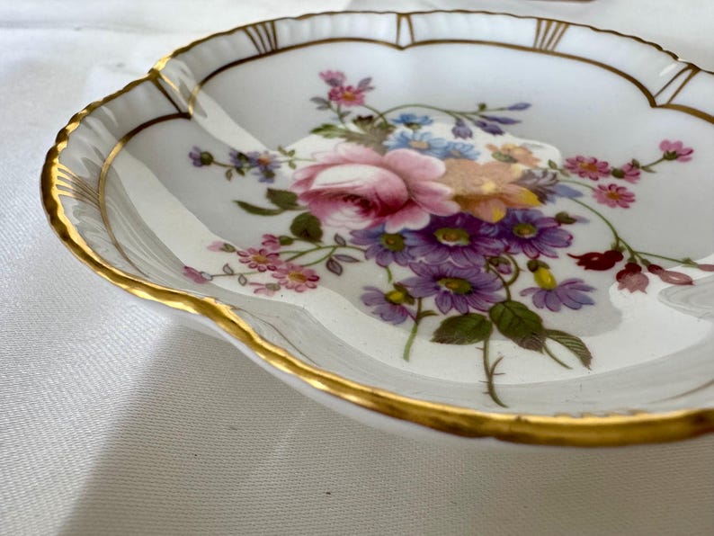 May include: A white porcelain dish with a scalloped edge and gold trim. The dish is decorated with a floral arrangement of pink roses, purple daisies, and other colorful flowers. A decorative piece for serving or display.