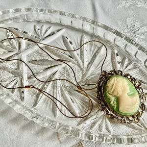 May include: A vintage cameo pendant necklace. The oval pendant features a cream-colored profile against a light green background, set in a gold-toned filigree frame. The necklace is displayed on a clear, textured glass dish.