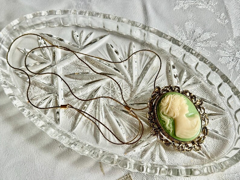 May include: A vintage cameo pendant necklace. The oval pendant features a cream-colored profile against a light green background, set in a gold-toned filigree frame. The necklace is displayed on a clear, textured glass dish.