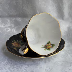 Pode incluir: Um conjunto de ch&aacute;vena de ch&aacute; e pires em porcelana com exterior preto e rebordo dourado. O interior da ch&aacute;vena branca e a superf&iacute;cie do pires est&atilde;o decorados com um motivo de bot&atilde;o de rosa amarelo. A ch&aacute;vena tem rebordo recortado e asa dourada.