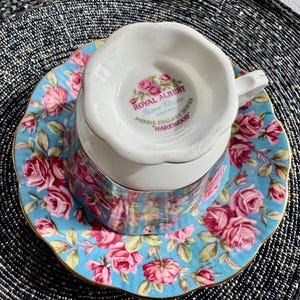K&ouml;nnte beinhalten: Eine wei&szlig;e Teetasse und Untertasse mit einem blauen und rosafarbenen Blumenmuster. Die Teetasse tr&auml;gt die Aufschrift "Royal Albert Bone China" und "Merrie England Series "Harewood" auf der Unterseite.
