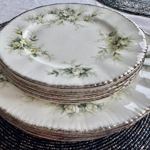 May include: A set of six white ceramic plates with a delicate floral design featuring white roses and green leaves. The plates have a gold rim and are stacked on top of each other.