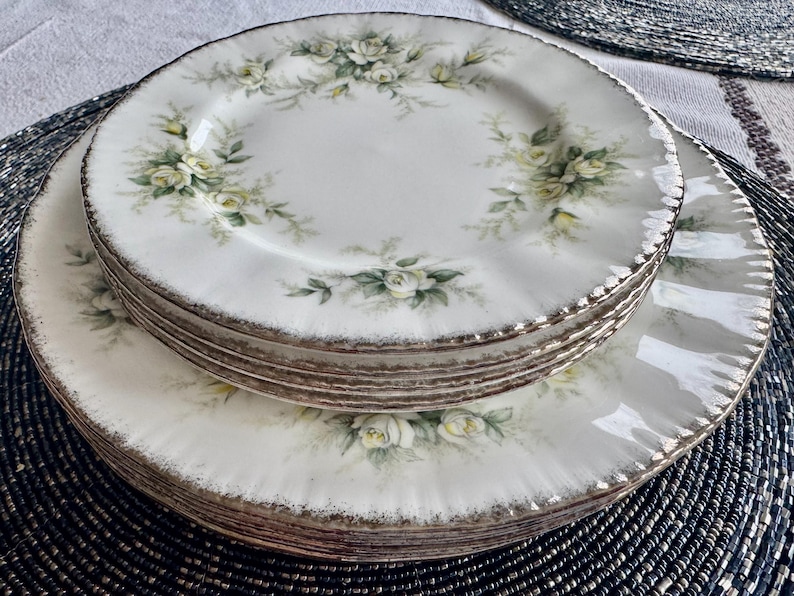 May include: A set of six white ceramic plates with a delicate floral design featuring white roses and green leaves. The plates have a gold rim and are stacked on top of each other.