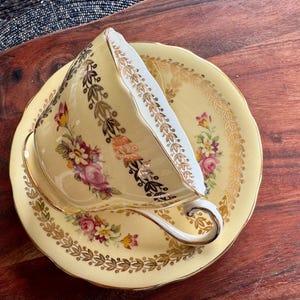 May include: A pale yellow porcelain teacup and saucer set featuring a delicate floral pattern with pink, purple, and yellow flowers. The rim of the cup and saucer is decorated with intricate gold leaf filigree and scalloped edges.