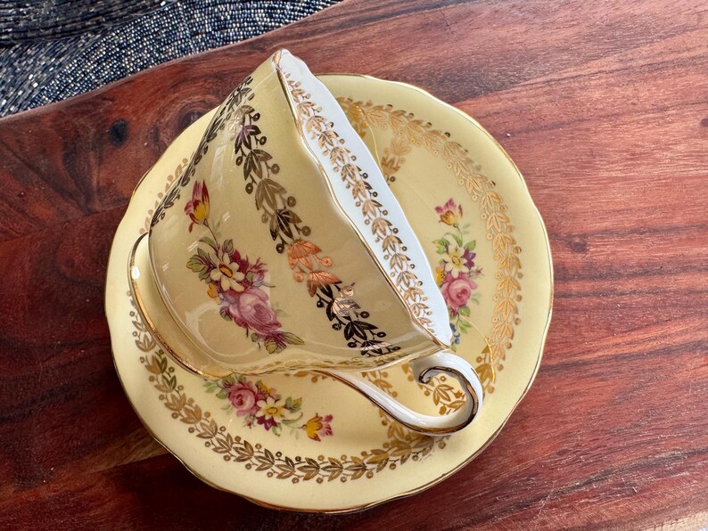 May include: A pale yellow porcelain teacup and saucer set featuring a delicate floral pattern with pink, purple, and yellow flowers. The rim of the cup and saucer is decorated with intricate gold leaf filigree and scalloped edges.