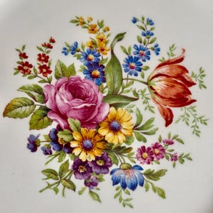 May include: White ceramic plate with a floral design. The design features a bouquet of flowers in shades of pink, red, yellow, blue, and purple, with green leaves. The plate has a gold rim.