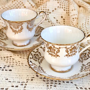 May include: Two white porcelain teacups and matching saucers featuring intricate gold filigree patterns. Each cup has a curved handle and a pedestal base, with gold trim along the rims and edges of the saucers, showcasing detailed floral motifs.
