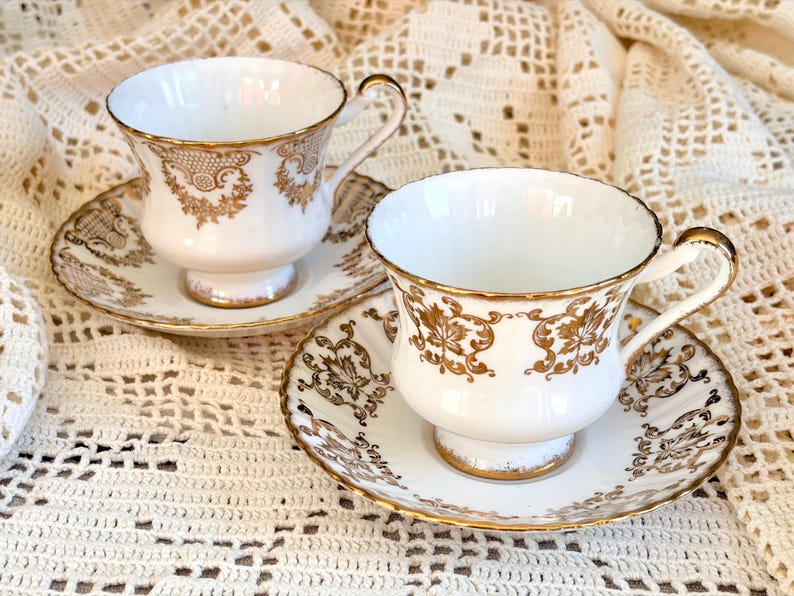 May include: Two white porcelain teacups and matching saucers featuring intricate gold filigree patterns. Each cup has a curved handle and a pedestal base, with gold trim along the rims and edges of the saucers, showcasing detailed floral motifs.