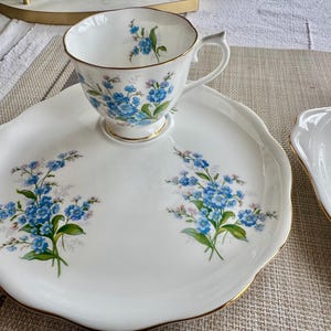 May include: A white porcelain teacup and saucer set with a floral design featuring blue forget-me-not flowers. The saucer has a scalloped edge and a gold trim.