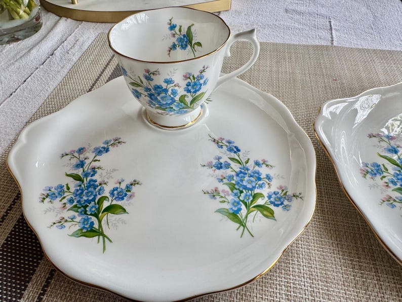 May include: A white porcelain teacup and saucer set with a floral design featuring blue forget-me-not flowers. The saucer has a scalloped edge and a gold trim.