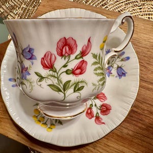 May include: A white teacup and saucer with a floral pattern featuring pink, blue, and green flowers. The cup has a gold rim and a delicate handle.