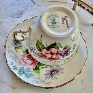 Pu&ograve; includere: Un set vintage di tazza e piattino da t&egrave;. La tazza e il piattino in porcellana bianca sono decorate con un motivo floreale rosa, viola e blu. I bordi sono rifiniti in oro. La tazza ha un manico curvo.