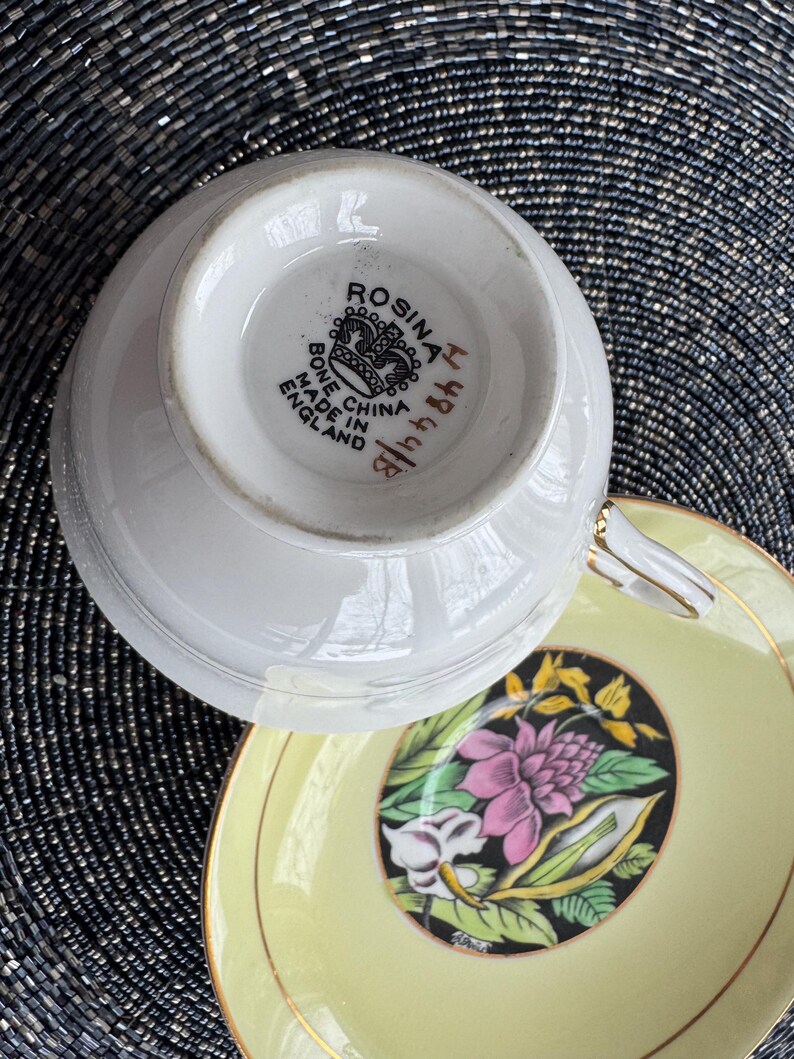 May include: A vintage teacup and saucer set with a floral design. The saucer is yellow with a black floral pattern. The teacup is white with a gold rim and the words "Rosina  Bone China Made in England" on the bottom.