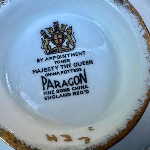 May include: The underside of a white fine bone china piece featuring a gold-trimmed rim. The center displays a black printed royal crest and text reading BY APPOINTMENT TO HER MAJESTY THE QUEEN CHINA POTTERS PARAGON FINE BONE CHINA ENGLAND REG'D.