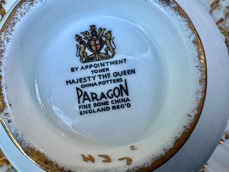 May include: The underside of a white fine bone china piece featuring a gold-trimmed rim. The center displays a black printed royal crest and text reading BY APPOINTMENT TO HER MAJESTY THE QUEEN CHINA POTTERS PARAGON FINE BONE CHINA ENGLAND REG'D.