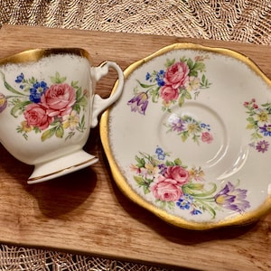 May include: A white teacup and saucer with a floral pattern in shades of pink, purple, and yellow. The edges of the cup and saucer are trimmed with gold.