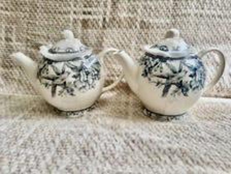 May include: Two cream-colored ceramic teapots featuring a dark blue transferware pattern of birds perched on flowering branches. Each teapot includes a matching lid with a small knob handle, a curved spout, and a rounded handle on the side.