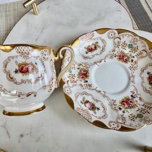 K&ouml;nnte beinhalten: Ein Vintage-Teebecher- und Untertassen-Set. Die wei&szlig;e Porzellantasse und Untertasse weisen ein Blumendesign mit rosa und blauen Blumen auf, das mit goldenen Verzierungen akzentuiert ist. Die Tasse hat einen Henkel und steht neben der passenden Untertasse.