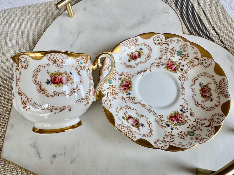 K&ouml;nnte beinhalten: Ein Vintage-Teebecher- und Untertassen-Set. Die wei&szlig;e Porzellantasse und Untertasse weisen ein Blumendesign mit rosa und blauen Blumen auf, das mit goldenen Verzierungen akzentuiert ist. Die Tasse hat einen Henkel und steht neben der passenden Untertasse.