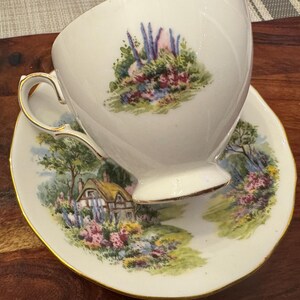 May include: A white teacup and saucer with a gold trim. The teacup and saucer feature a floral design with a cottage in the background.