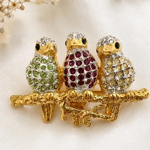 May include: A gold-toned brooch featuring three bejeweled birds perched on a branch. The birds are adorned with green, red, and yellow crystals, with black eyes. The branch has a detailed, textured design.