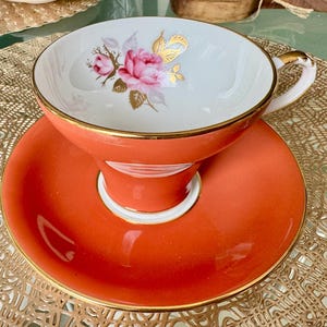 May include: A vintage teacup and saucer set with a floral design. The cup and saucer are both a vibrant orange color with a gold trim. The cup features a delicate floral design with pink roses and gold accents.