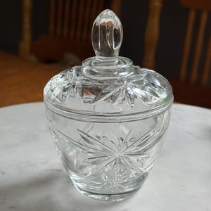 May include: A clear glass sugar bowl with a matching lid featuring a pointed finial. The body and lid are decorated with a pressed starburst pattern. The glass is transparent and reflects light, showcasing the intricate geometric star designs.