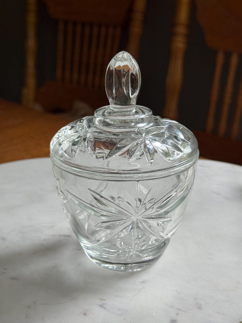 May include: A clear glass sugar bowl with a matching lid featuring a pointed finial. The body and lid are decorated with a pressed starburst pattern. The glass is transparent and reflects light, showcasing the intricate geometric star designs.