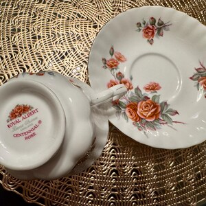 Pode incluir: Um conjunto de ch&aacute;vena e pires branco com um design floral. A ch&aacute;vena de ch&aacute; tem as palavras "Royal Albert Bone China Centennial Rose" na parte inferior.