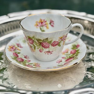 May include: A white teacup and saucer set with a floral design featuring pink and yellow flowers and green leaves. The set has gold trim and sits on a silver tray.