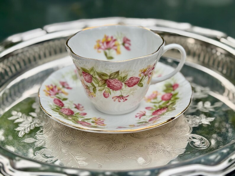 May include: A white teacup and saucer set with a floral design featuring pink and yellow flowers and green leaves. The set has gold trim and sits on a silver tray.