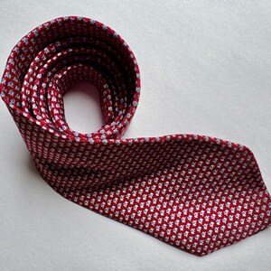 Puede incluir: Una corbata roja con un patr&oacute;n repetitivo de peque&ntilde;os motivos florales en blanco y azul claro. La corbata est&aacute; enrollada en la parte superior y se extiende hacia abajo, mostrando la textura de la tela y la disposici&oacute;n de las flores.