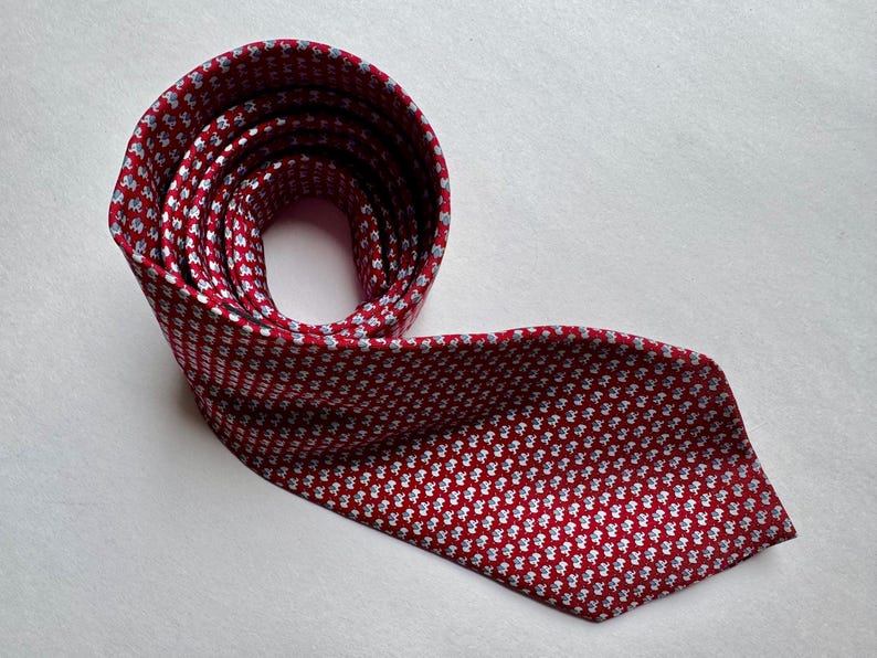 Puede incluir: Una corbata roja con un patr&oacute;n repetitivo de peque&ntilde;os motivos florales en blanco y azul claro. La corbata est&aacute; enrollada en la parte superior y se extiende hacia abajo, mostrando la textura de la tela y la disposici&oacute;n de las flores.