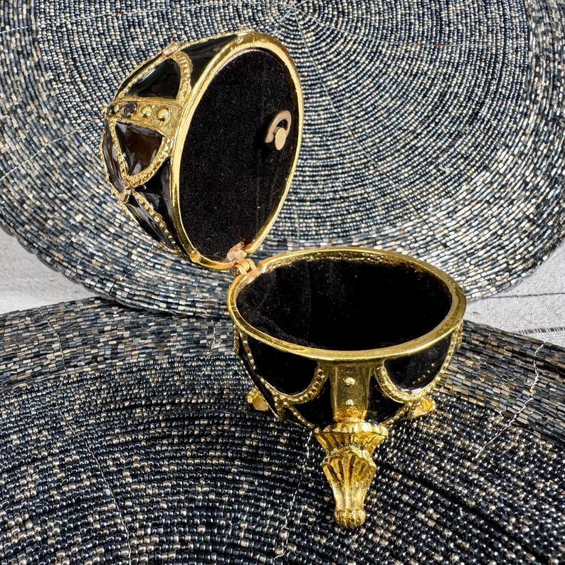 May include: A black and gold egg-shaped trinket box with a hinged lid and three gold feet. The box is open and the inside is lined with black velvet.