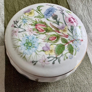 Op de afbeelding: Een ronde, witte porseleinen sieradendoos met een bloemenontwerp. Het deksel heeft een boeket van kleurrijke bloemen, waaronder roze, blauwe en paarse bloemen, met groene bladeren. De doos heeft een gouden rand en een geschulpte rand.