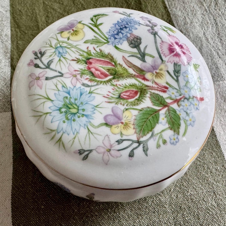 Op de afbeelding: Een ronde, witte porseleinen sieradendoos met een bloemenontwerp. Het deksel heeft een boeket van kleurrijke bloemen, waaronder roze, blauwe en paarse bloemen, met groene bladeren. De doos heeft een gouden rand en een geschulpte rand.