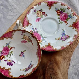 May include: A vintage teacup and saucer set. The white porcelain cup and saucer feature a pink and gold trim with a floral design. The cup has a handle and the saucer is round. The set is placed on a wooden surface.