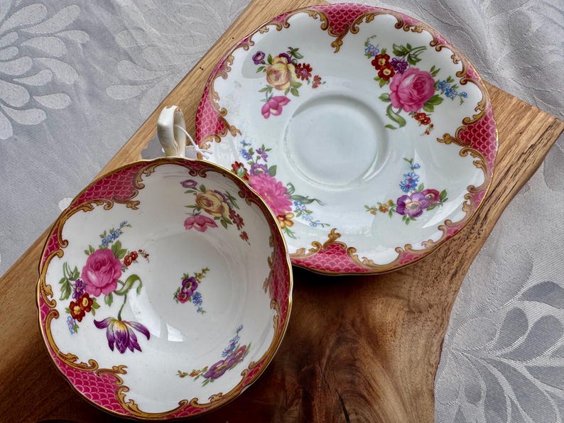 May include: A vintage teacup and saucer set. The white porcelain cup and saucer feature a pink and gold trim with a floral design. The cup has a handle and the saucer is round. The set is placed on a wooden surface.